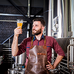 ERP for beverage manufacturing used in modern brewery Worker in modern brewery looks at a glass of beer produced using ERP for beverage manufacturing